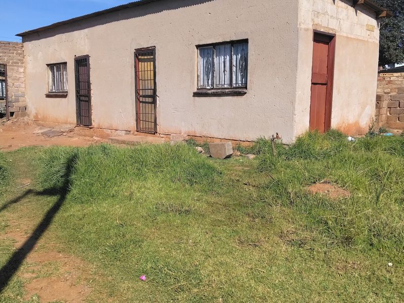 A single-story residential structure featuring a corrugated metal roof, security gates, and exterior plaster finish, set on a grassed plot.