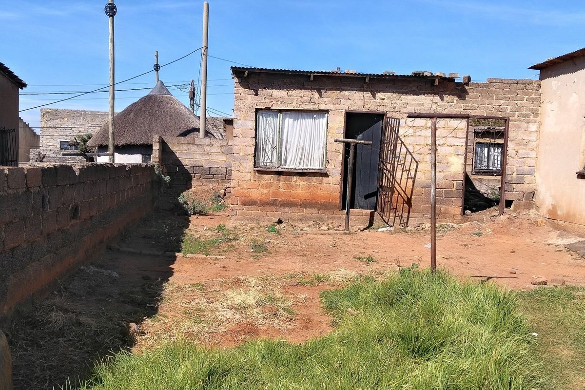 Rear view of a modest brick house featuring a small yard, metal security gate, and corrugated metal roofing.