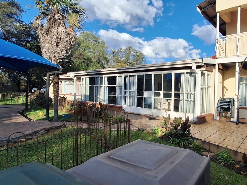 Rear view of the property featuring a sunroom, tiled patio area, upper balcony, and a shaded outdoor space with established garden surroundings.