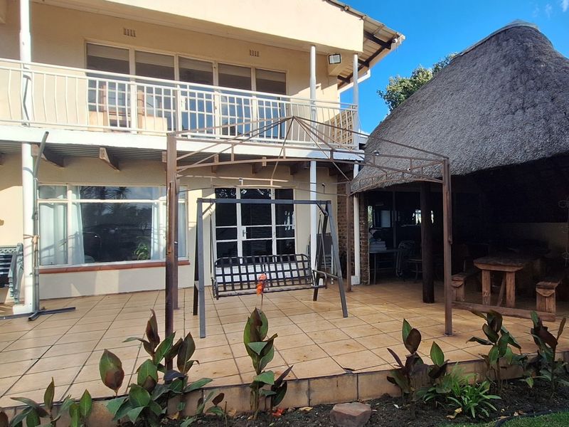 Rear view of the property featuring a tiled patio area, a traditional thatched lapa, and an upper-level balcony with metal railings.