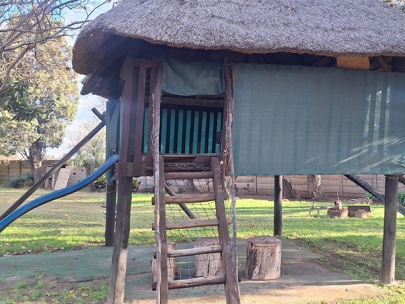A rustic outdoor play structure featuring a thatched roof, wooden support posts, a slide, and a climbing ladder set within a garden area.