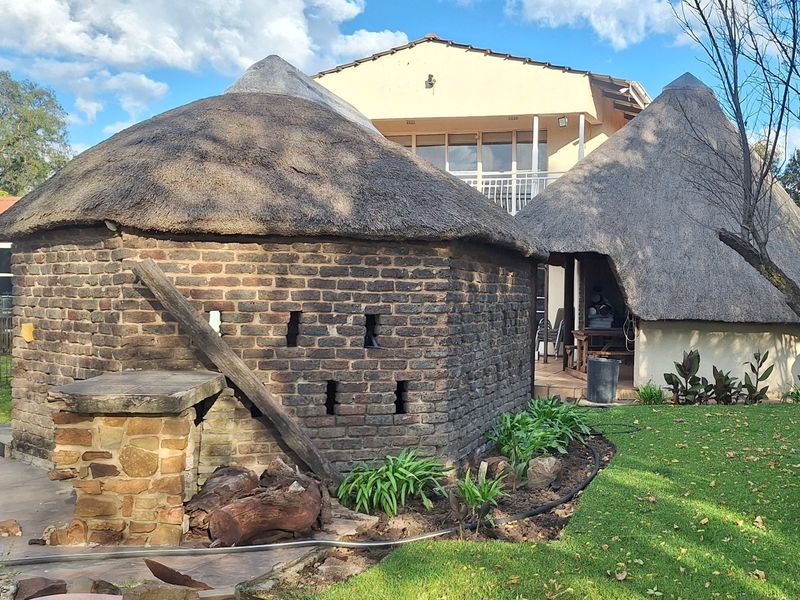 Rear garden view featuring traditional thatch-roofed structures, exposed brickwork, and a stone masonry braai area set within a landscaped lawn.