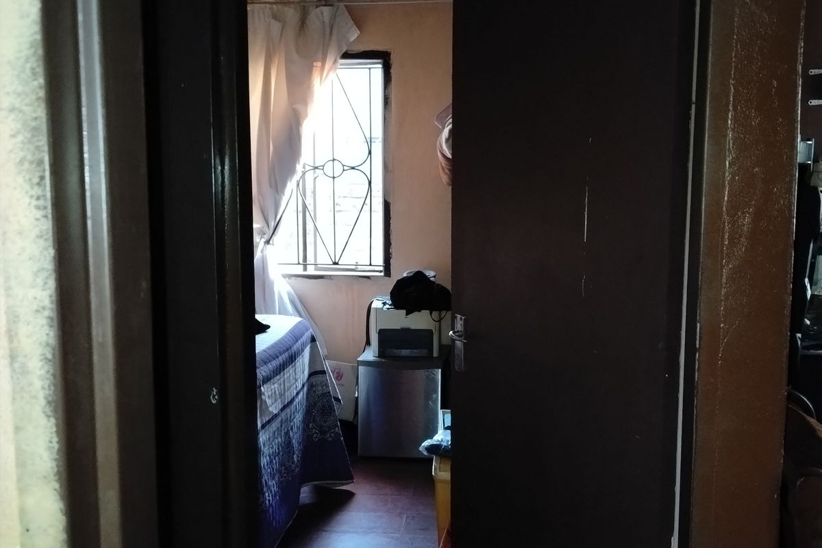 A view into a bedroom featuring tiled flooring and a security gate on the window, showing signs of wear and dated finishes.