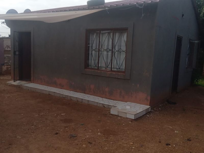 A single-story residential dwelling featuring a metal roof, security bars on windows, and a tiled entrance step, set on an unpaved ground surface.
