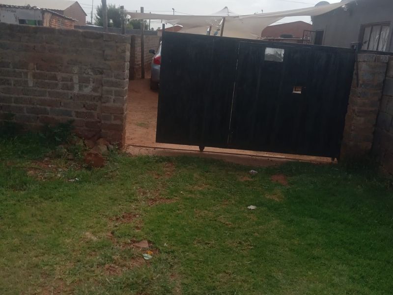 Front exterior view showing a brick boundary wall, metal driveway gate, and a shade sail over the entrance area.