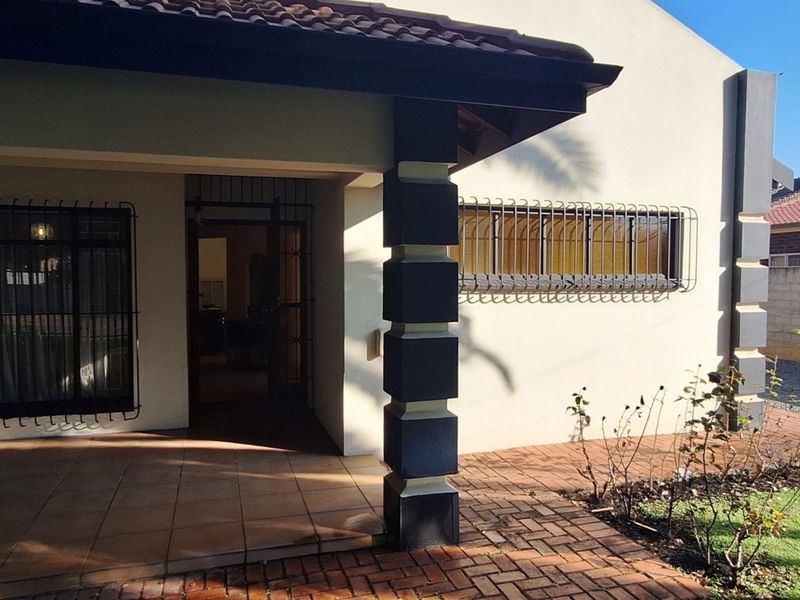 A covered patio area featuring tiled flooring, security bars on windows, and adjacent brick-paved garden pathways.