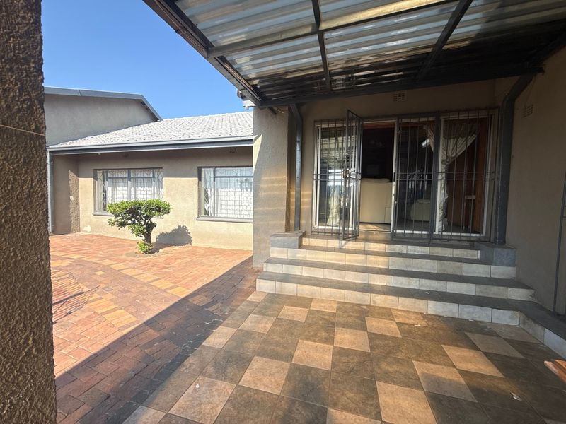 A covered patio area featuring tiled flooring and concrete steps, leading into the home through a secure metal gate, overlooking a brick-paved courtyard.