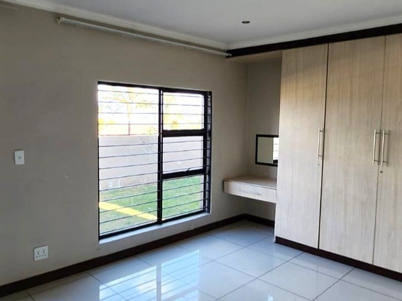 A bright, empty bedroom featuring neutral tiled flooring, extensive built-in cupboards, a integrated vanity unit, and security bars on the windows.
