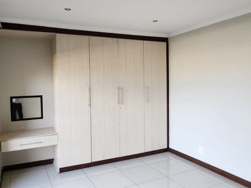 A clean, empty bedroom featuring light wood-finish built-in cupboards, a matching vanity unit, tiled flooring, and security bars on the window.
