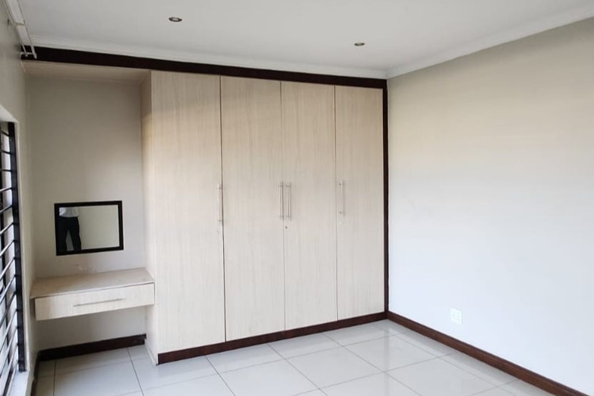 A clean, empty bedroom featuring light wood-finish built-in cupboards, a matching vanity unit, tiled flooring, and security bars on the window.