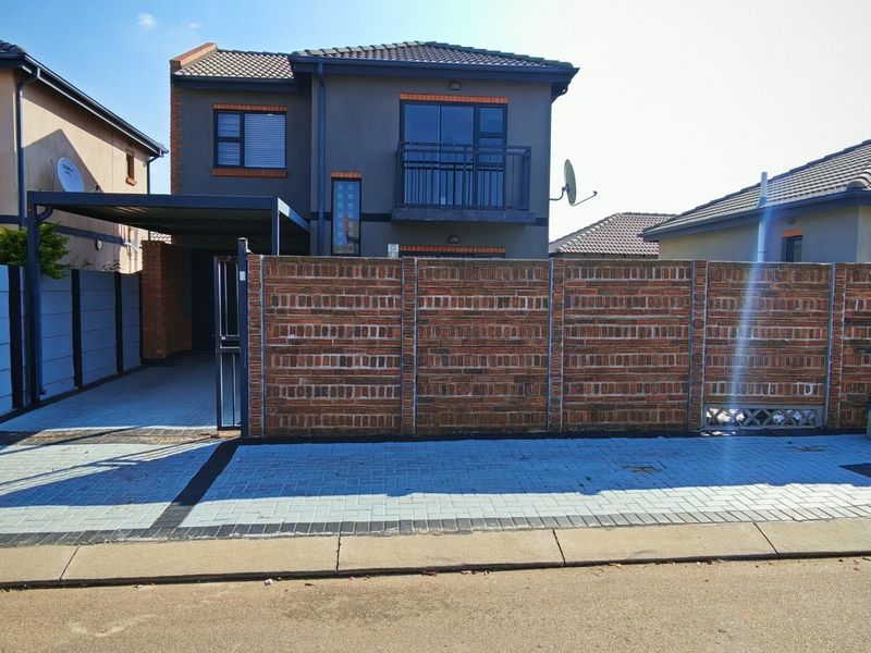 Front view of a two-story residential house featuring a paved driveway, carport, and secure brick boundary wall with a metal gate.