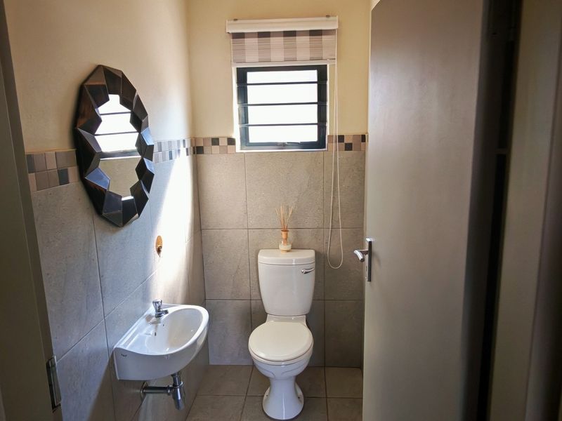 A compact guest bathroom featuring neutral wall tiles, a wall-mounted basin, and a standard toilet with a louvered window for ventilation.