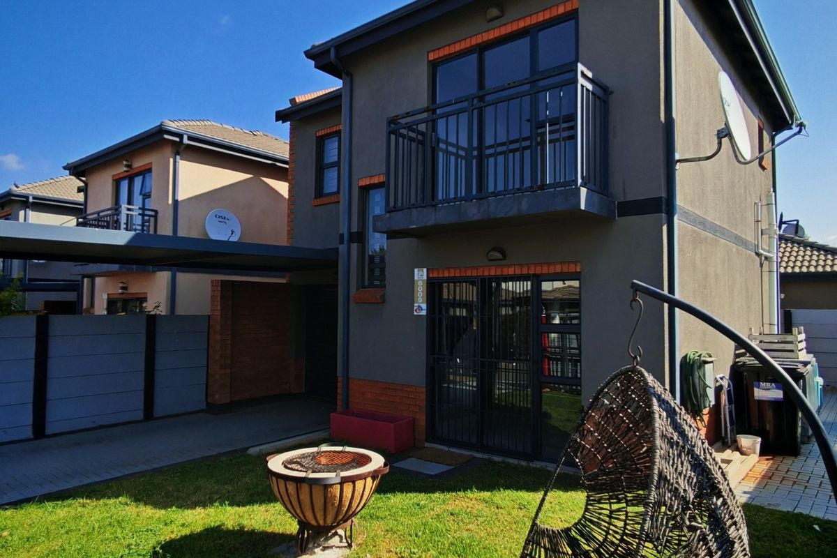 Rear view of a modern double-story townhouse featuring a private garden, paved driveway, carport, and an upper-level balcony with metal railings.