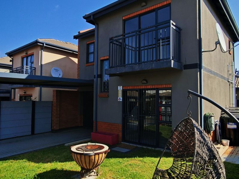 Rear view of a modern double-story townhouse featuring a private garden, paved driveway, carport, and an upper-level balcony with metal railings.