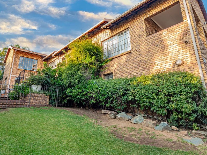 Rear view of a double-storey face brick house featuring a balcony, steel-framed windows, and a landscaped garden with a retaining wall.
