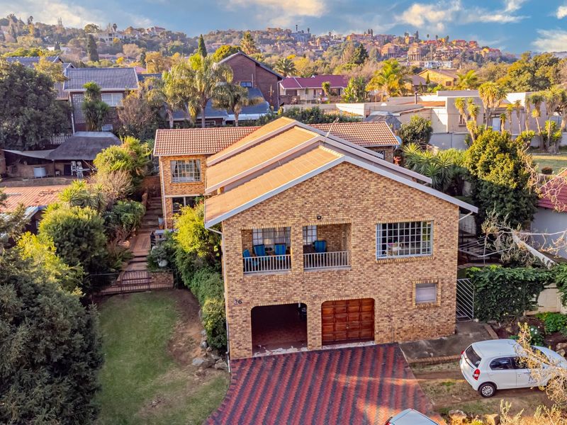 Aerial view of a double-story face brick house featuring a tiled roof, balcony, double garage, and a paved driveway set within a landscaped garden.