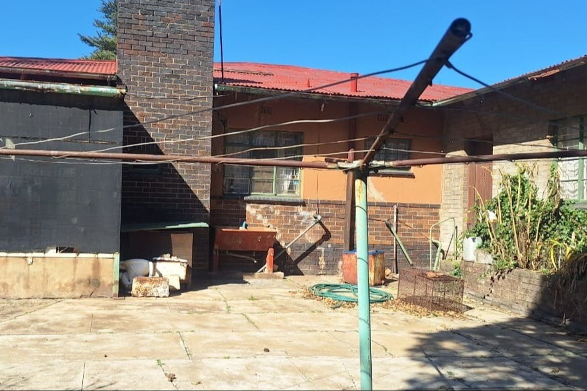 Rear exterior view of the property featuring a brick chimney, corrugated metal roofing, and a paved patio area requiring maintenance.