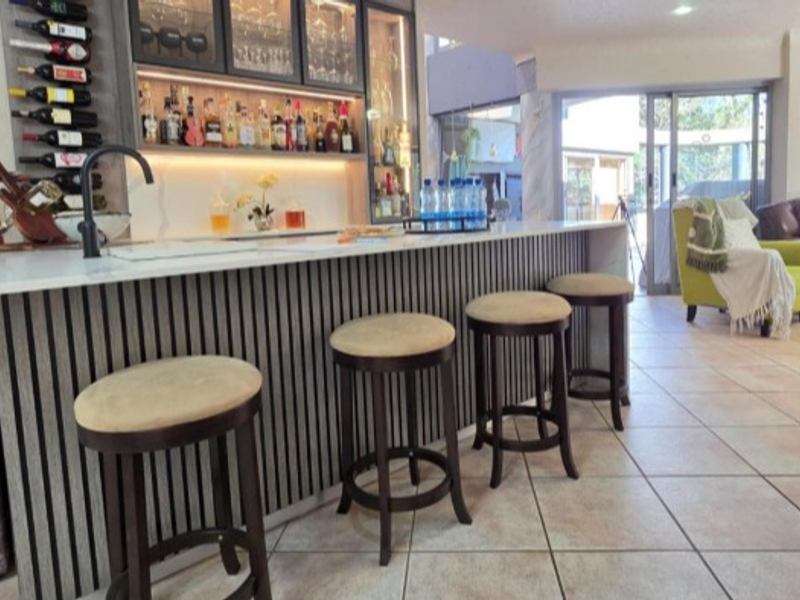A modern home bar area featuring a sleek slatted timber counter, integrated shelving with ambient lighting, and tiled flooring leading to an outdoor space.
