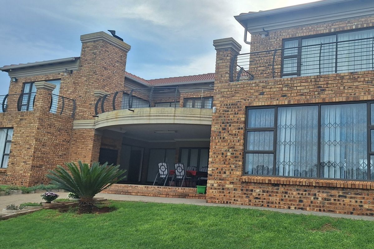 Rear view of a multi-story face brick residence featuring a covered patio, upper-level balcony with metal railings, and large aluminium-framed windows.