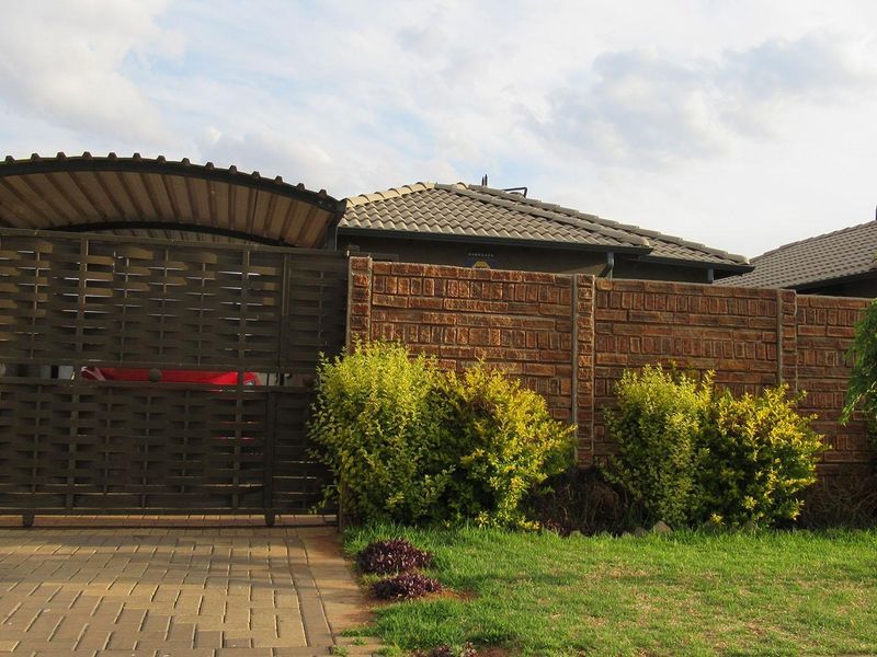 Front view of the property featuring a paved driveway, metal sliding gate, carport, and a brick boundary wall with established garden landscaping.