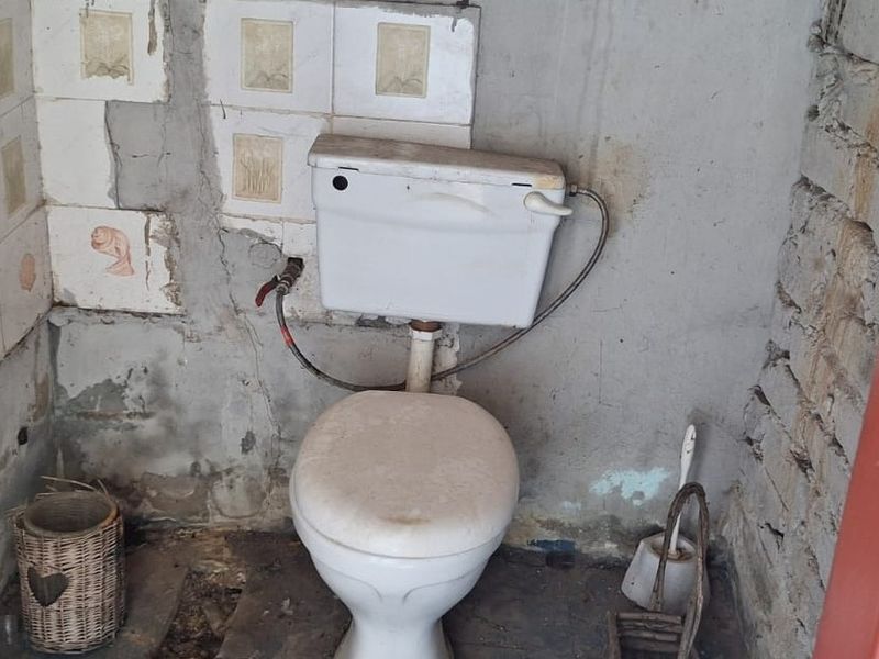 A small, unfinished bathroom featuring a standard toilet, exposed brickwork, and damaged wall plaster requiring significant renovation and repair.