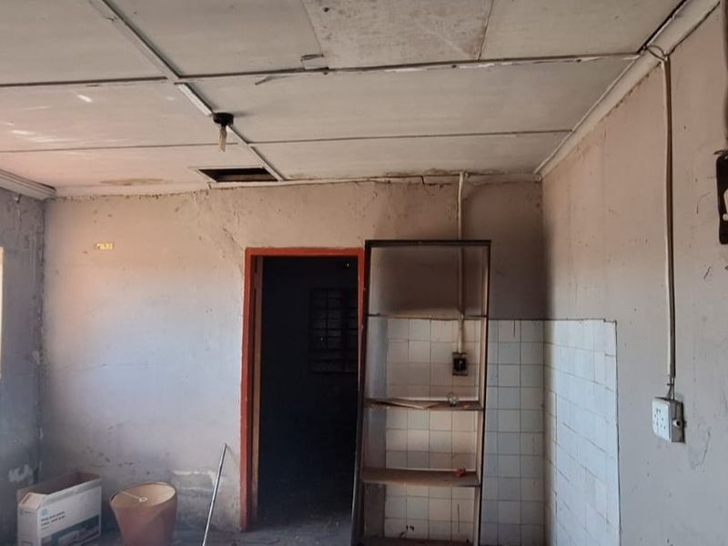 A dilapidated kitchen space requiring significant renovation, featuring exposed wiring, damaged ceiling tiles, and basic tiled splashback.