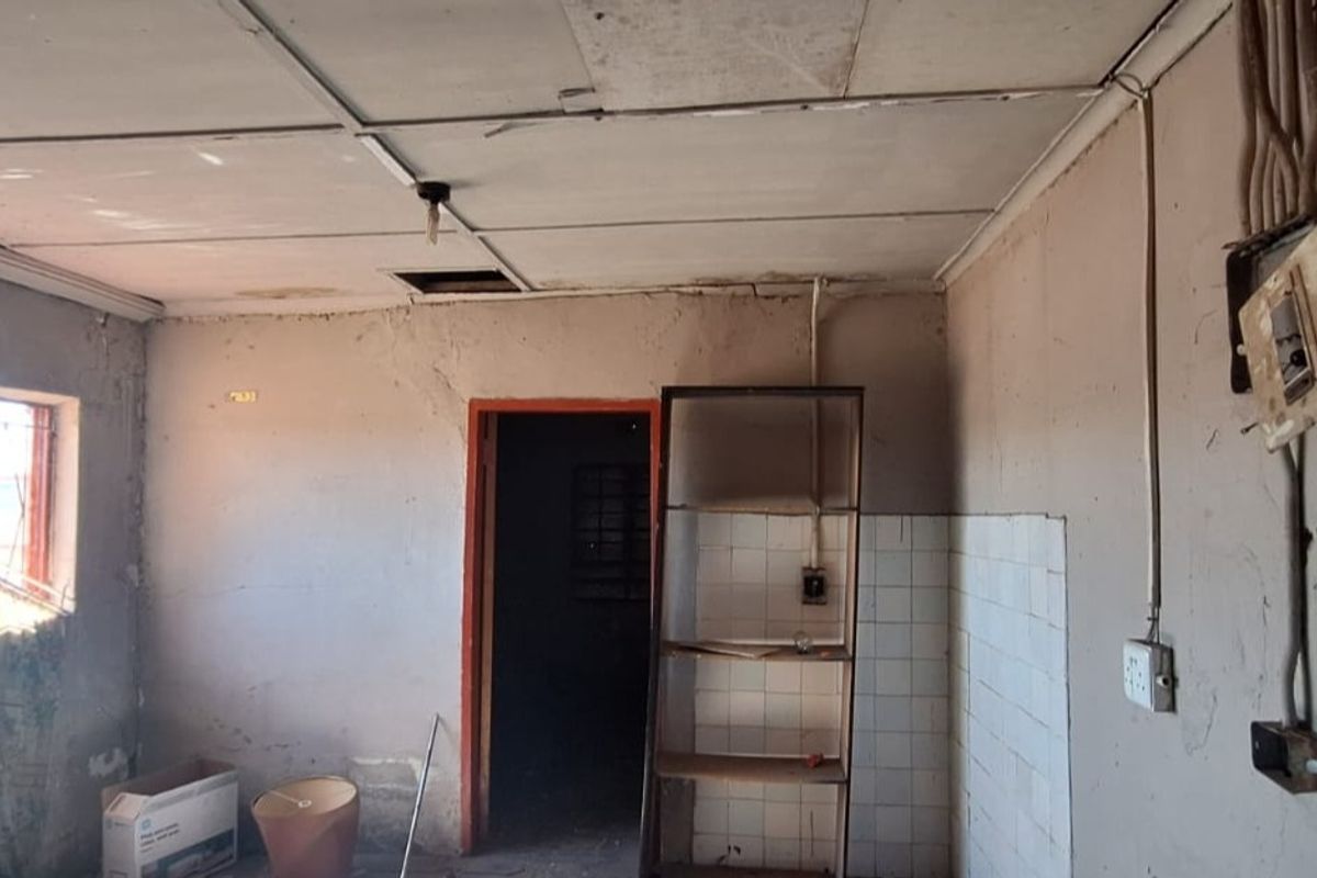 A dilapidated kitchen space requiring significant renovation, featuring exposed wiring, damaged ceiling tiles, and basic tiled splashback.