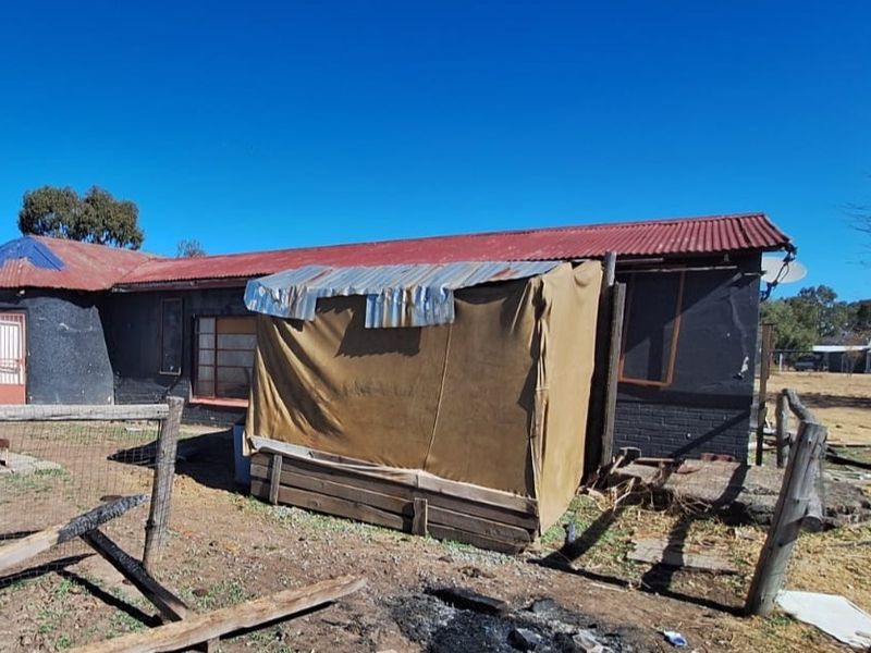 Rear view of the property showing brick masonry walls, corrugated iron roofing, and an attached makeshift structure in a rural setting.