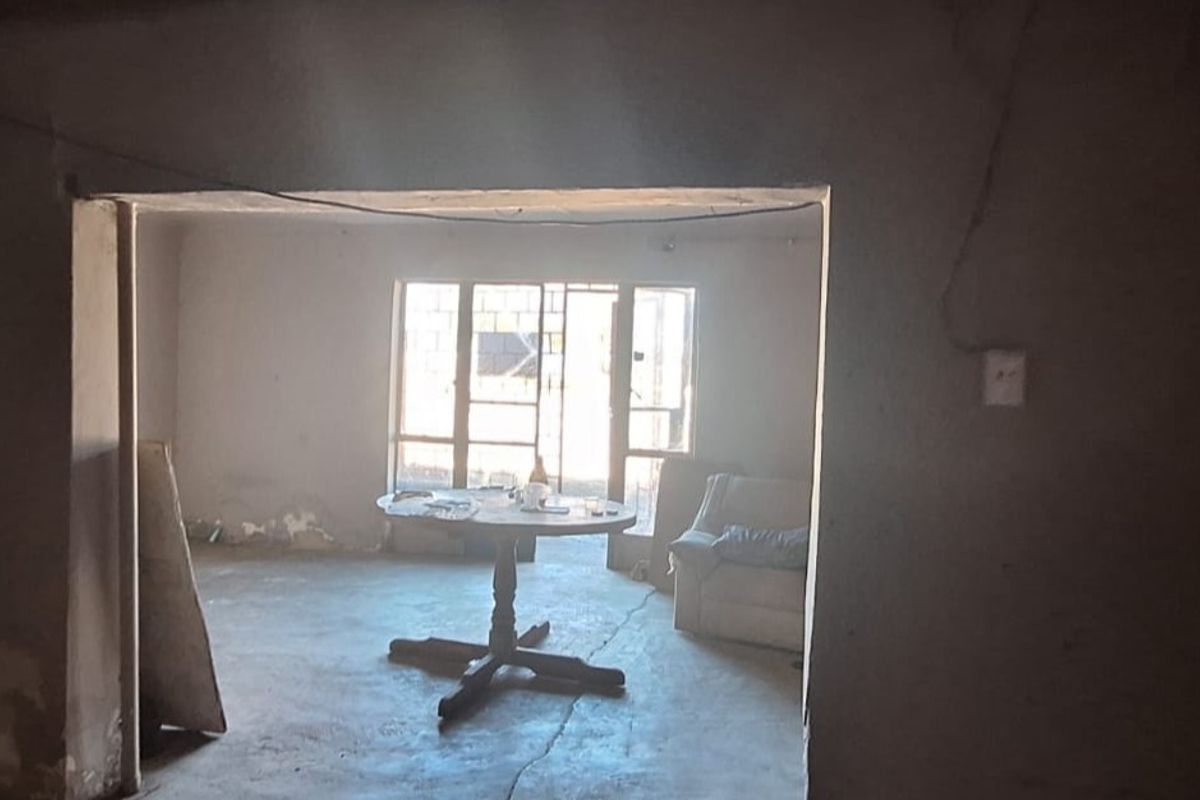 An interior view of a living area in poor condition, featuring concrete flooring, exposed wall plaster, and metal-framed windows leading to the exterior.