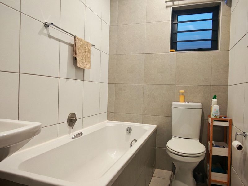 A functional bathroom featuring a bathtub, pedestal basin, and toilet, finished with neutral wall tiling and a louvered window for ventilation.