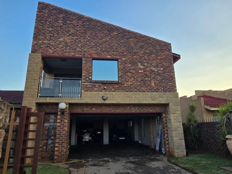 Front elevation of a double-storey face brick house featuring a double garage, upper-level balcony with glass balustrade, and paved driveway.