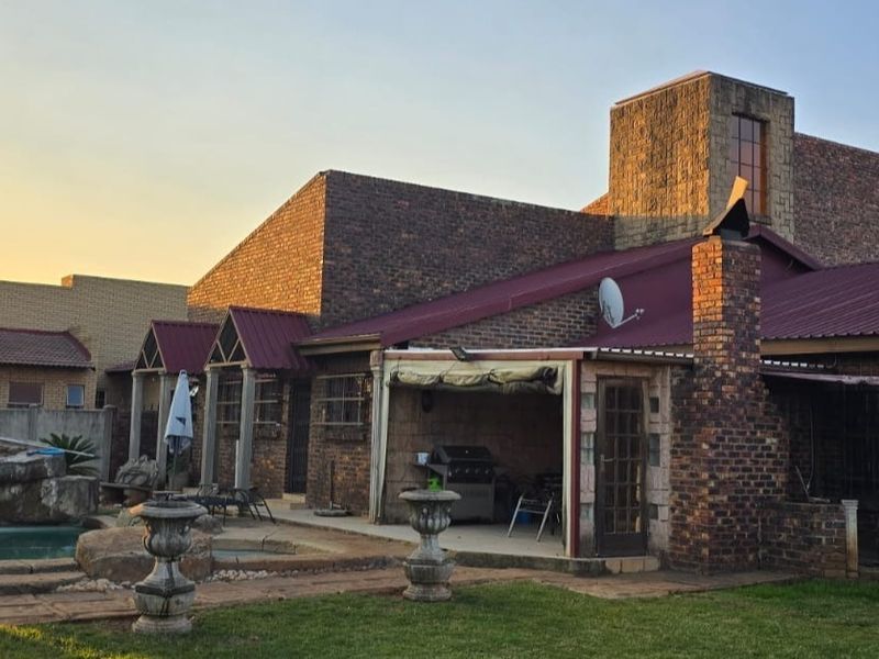 Rear view of a brick house featuring a covered patio, swimming pool, and metal roofing, set within a landscaped garden area.