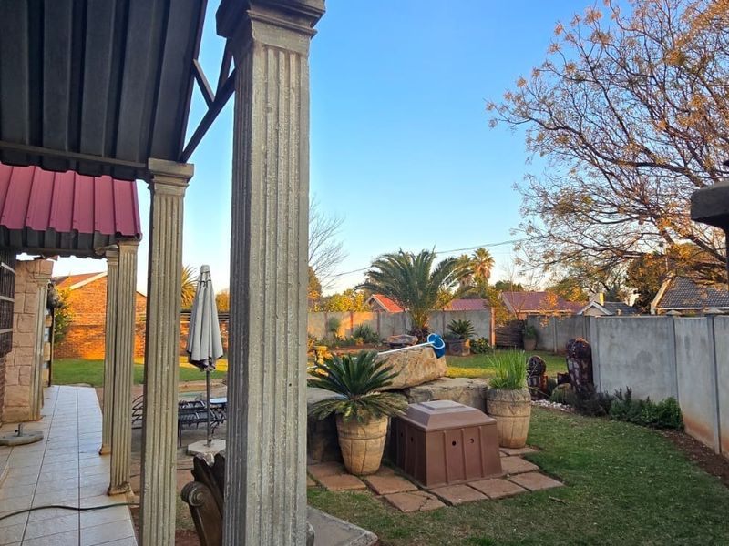 A rear garden view featuring a covered patio with concrete columns, tiled flooring, and a surrounding precast concrete boundary wall.