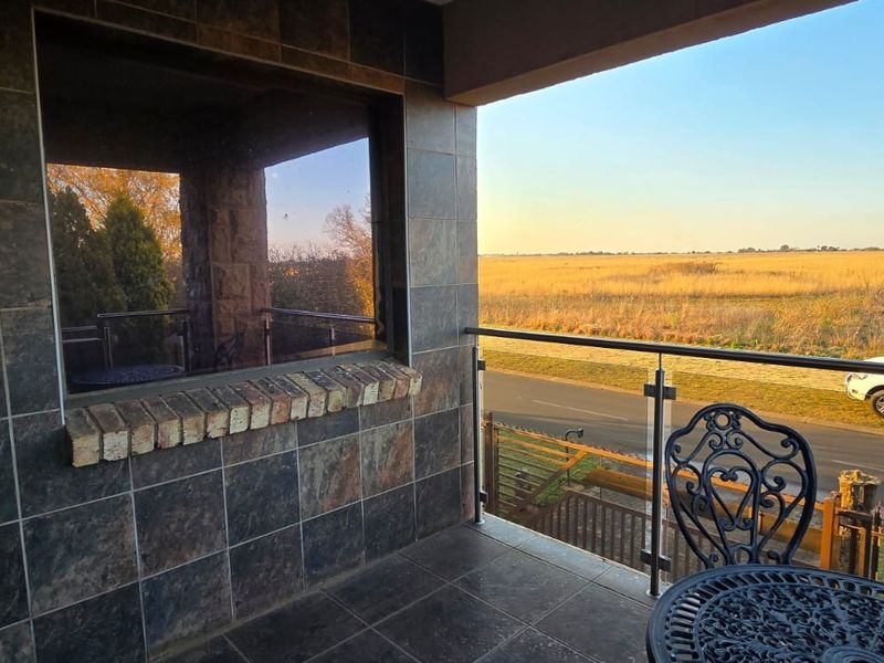 A covered balcony featuring slate wall and floor tiling, finished with a modern glass and stainless steel balustrade overlooking an open field.