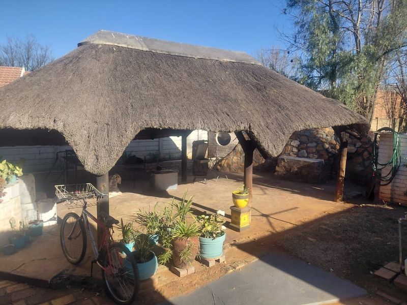 Outdoor thatched lapa area featuring timber support posts, stone walling, and brick paving, providing a rustic covered entertainment space in the garden.
