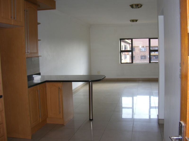 An open-plan living and kitchen area featuring tiled flooring, built-in wooden cabinetry with a breakfast bar, and large windows providing natural light.