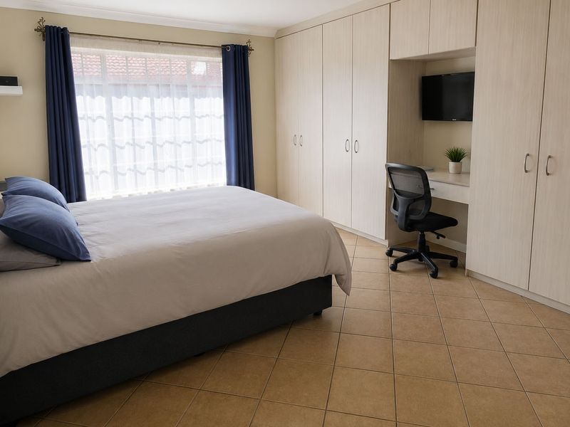 A well-maintained bedroom featuring extensive built-in cupboards, a integrated desk area, tiled flooring, and large windows with curtain rails.