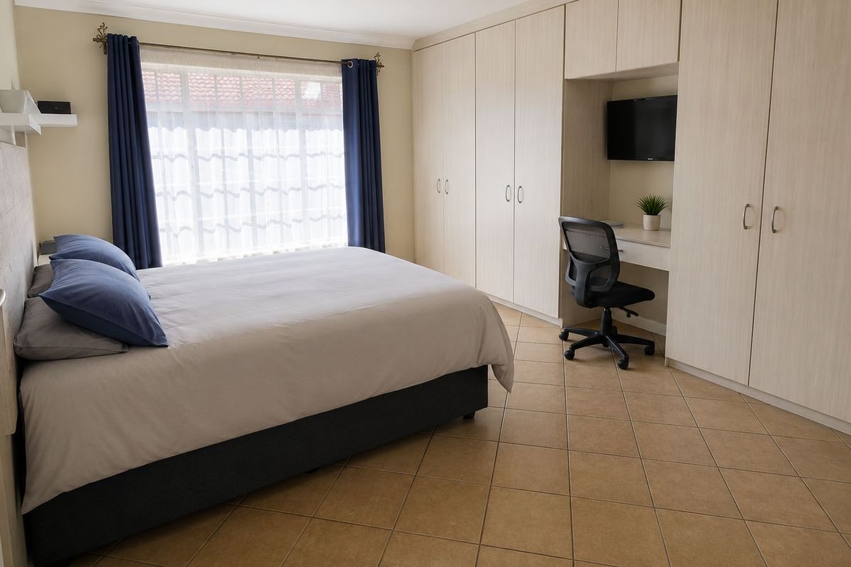A well-maintained bedroom featuring extensive built-in cupboards, a integrated desk area, tiled flooring, and large windows with curtain rails.