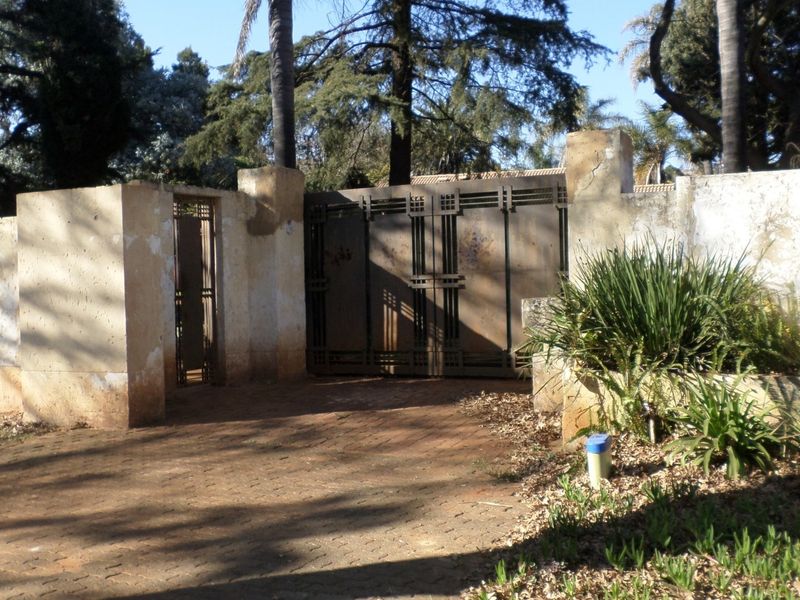 Front entrance featuring a paved driveway, metal security gates, and a perimeter boundary wall requiring maintenance.