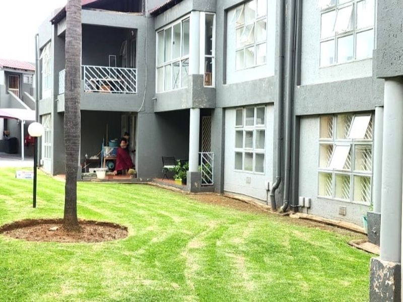 Rear view of a townhouse complex featuring a shared lawn area, covered patios, and multi-level residential units with security gates and casement windows.
