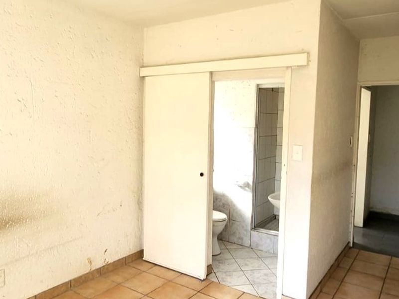 An empty bedroom featuring tiled flooring and an en-suite bathroom with a shower, toilet, and basin, requiring cosmetic updates to the wall finishes.