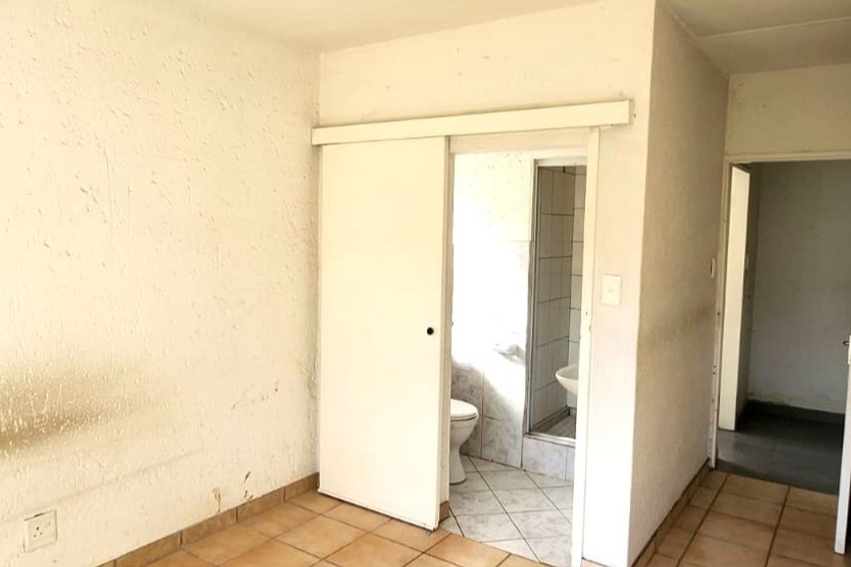 An empty bedroom featuring tiled flooring and an en-suite bathroom with a shower, toilet, and basin, requiring cosmetic updates to the wall finishes.