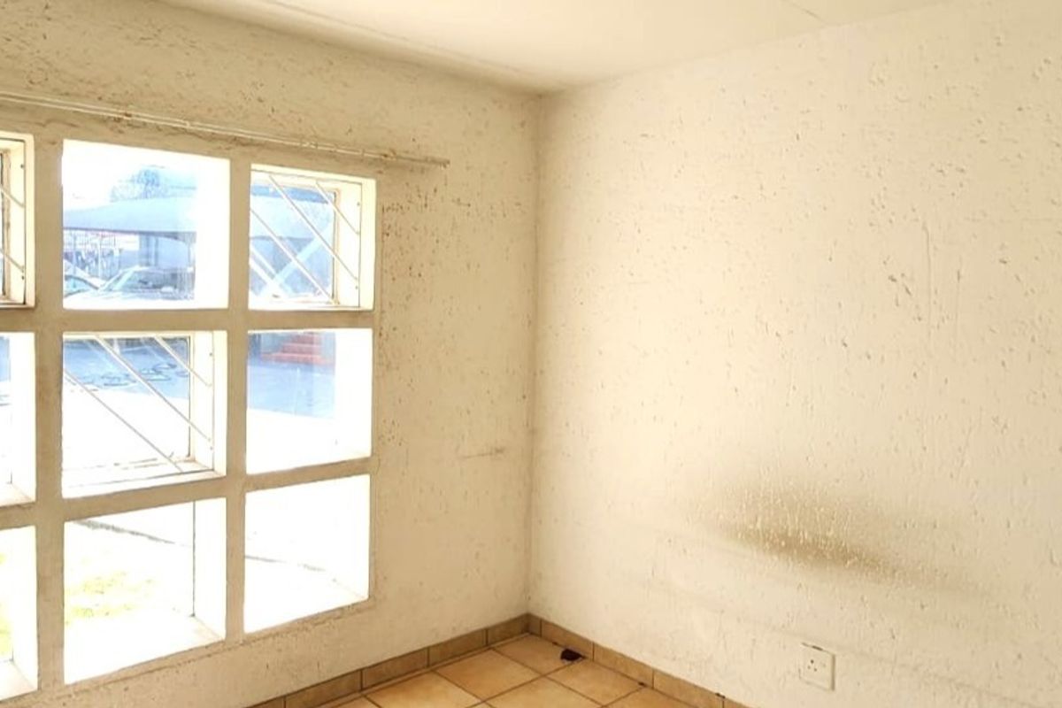 An empty living room featuring tiled flooring and large grid-pattern windows with security bars, requiring significant wall repairs and cosmetic updates.