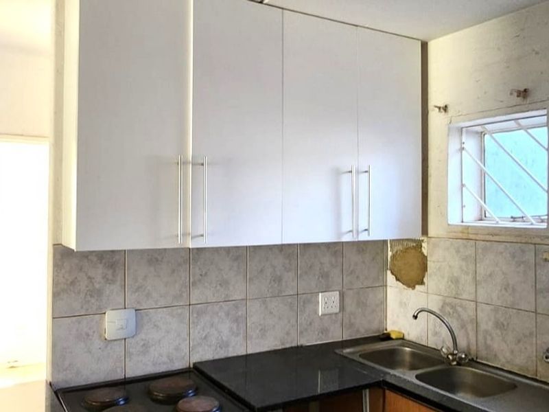 A compact kitchen featuring white cabinetry, a double sink, and an electric hob, currently requiring cosmetic repairs to the tiled backsplash and wall finishes.