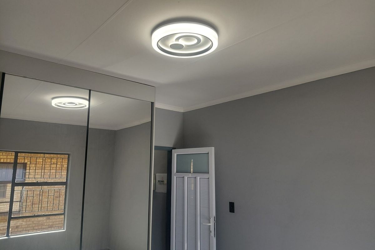 A clean, neutral bedroom featuring a large built-in mirrored wardrobe, modern LED ceiling lighting, and an aluminium window with security bars.