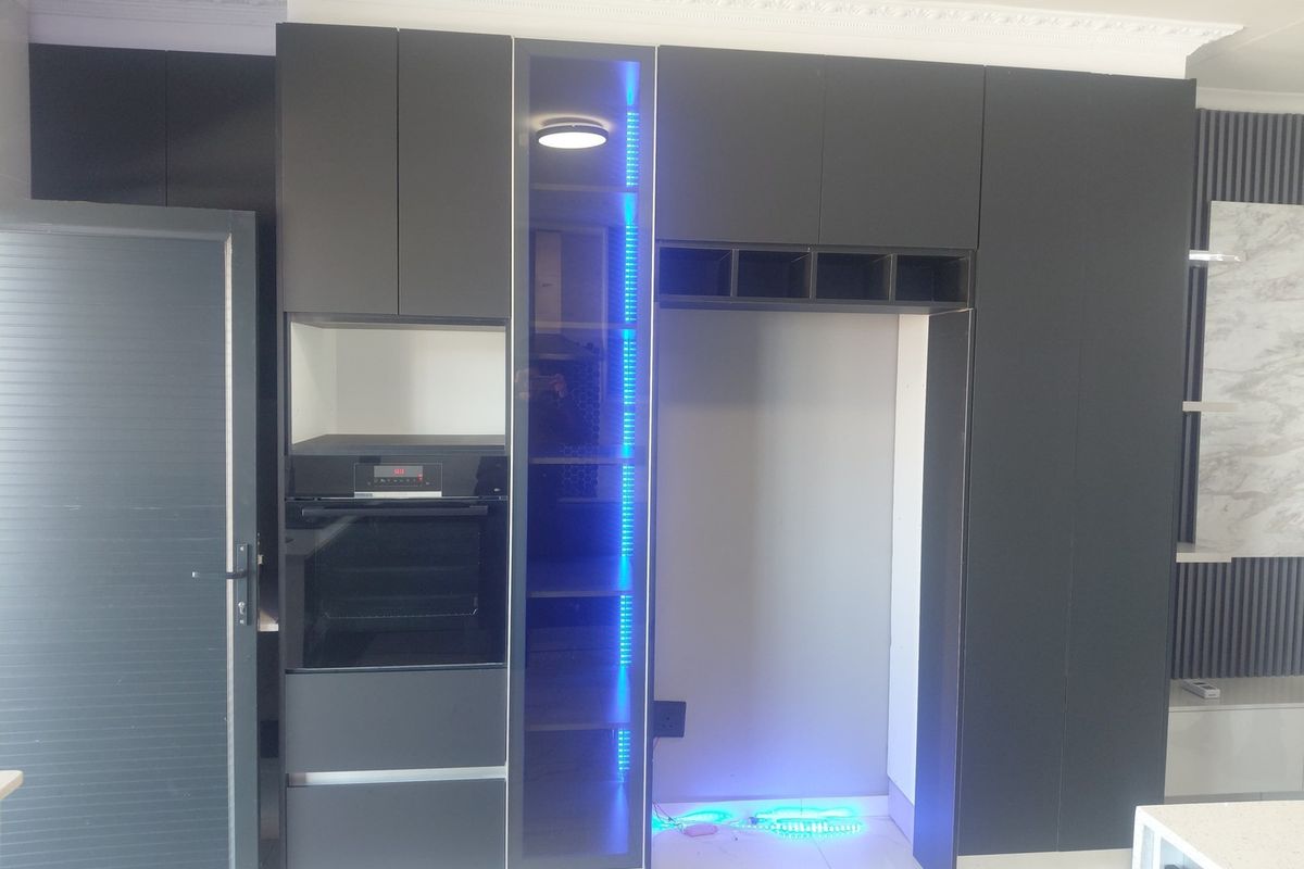 Modern kitchen wall featuring sleek dark cabinetry, a built-in oven, integrated display shelving with LED lighting, and a dedicated refrigerator alcove.