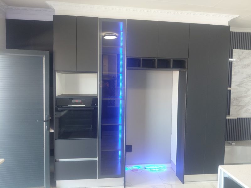 Modern kitchen wall featuring sleek dark cabinetry, a built-in oven, integrated display shelving with LED lighting, and a dedicated refrigerator alcove.