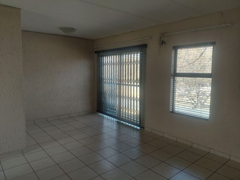 An empty living room featuring tiled flooring, a sliding glass door with security gate, and window blinds. The space requires minor cosmetic updates.
