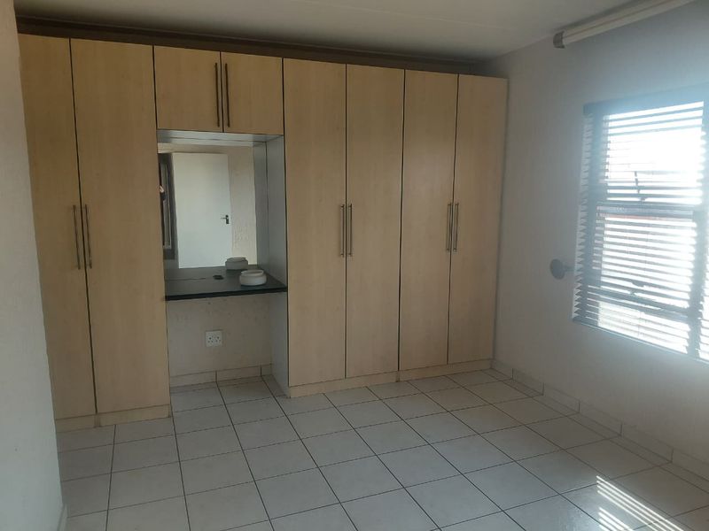 A bright, empty bedroom featuring extensive built-in cupboards with an integrated vanity station, tiled flooring, and venetian blinds.