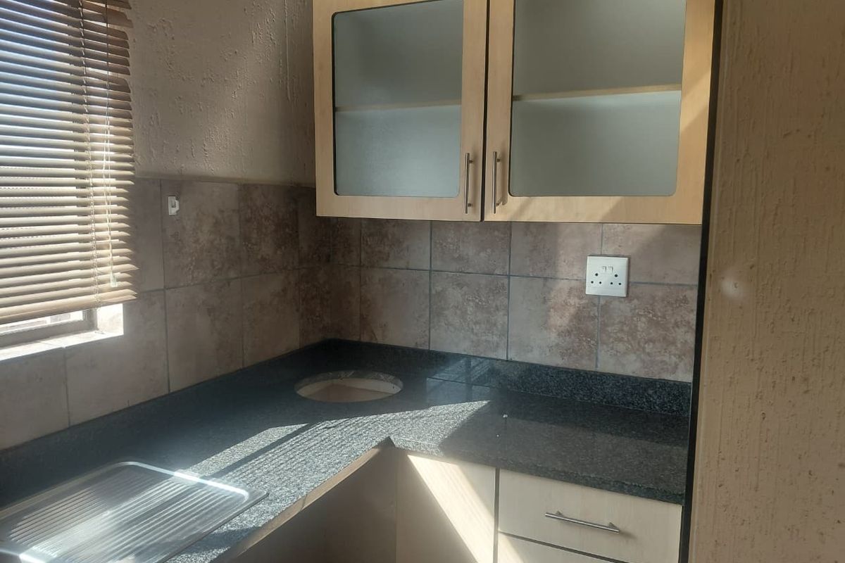A compact kitchen area featuring granite countertops, stainless steel sink, tiled backsplash, and light wood-finish cabinetry with glass-fronted upper units.